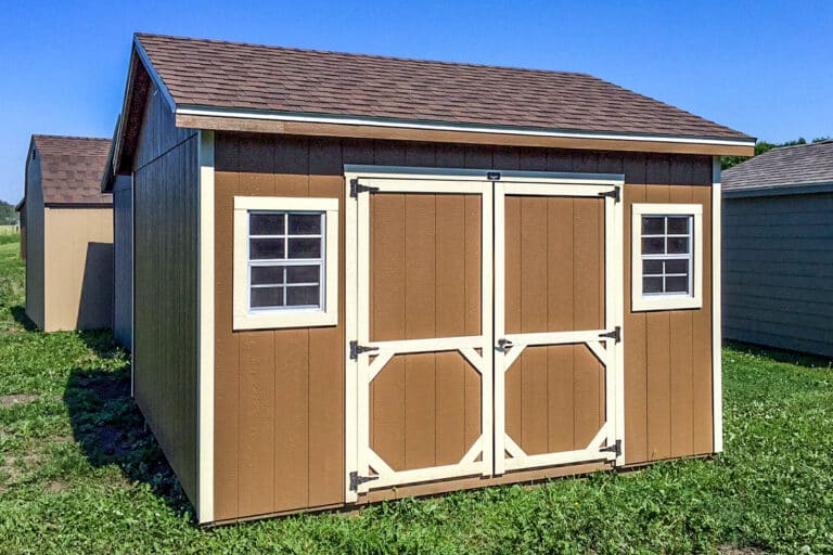 Brown Quaker-style shed with cream trim, double doors, and two front windows, standing on a grassy lot under a clear blue sky.