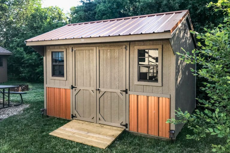 Brown shed with orange metal wainscoting, double doors, and two dark-trimmed windows, placed on grass with a wooden entry ramp.