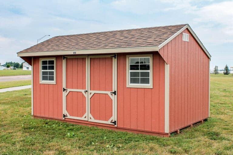 Red Quaker-style shed with tan double doors, matching trim, and two windows, set on a grassy lawn near a sidewalk.