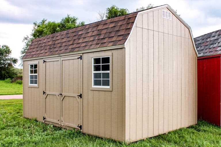 Beige gambrel-roof shed with double doors and two front-facing windows, set on a grassy lawn near a paved driveway.