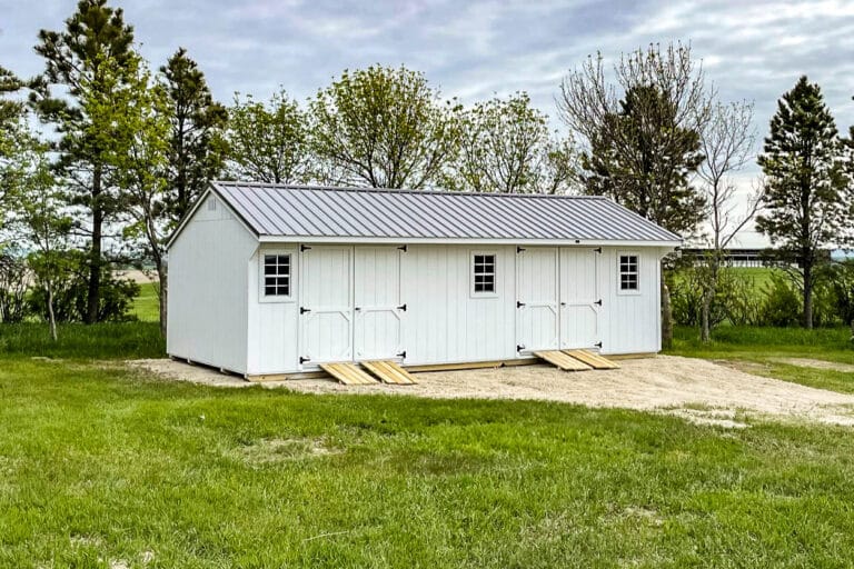 Long white Quaker-style shed with two sets of double doors, three windows, and a silver metal roof, positioned on a dirt pad in a grassy field near trees.