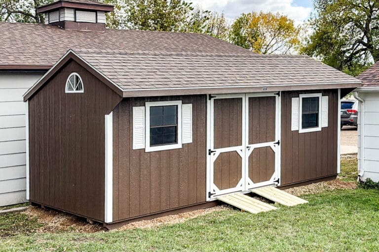 Long brown shed with white trim, dual window shutters, double doors with ramps, and a decorative gable window, placed beside a white building.