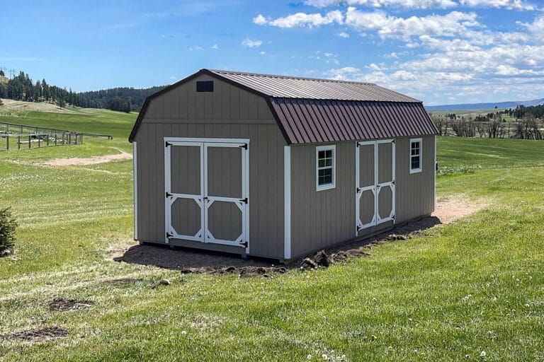 Light brown gambrel-roof shed with white-trimmed double doors and windows, set in a wide open grassy field under a blue sky.