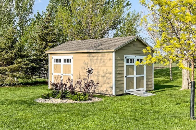 Tan shed with white trim and transom windows on double doors, surrounded by green grass, trees, and landscaped plants.