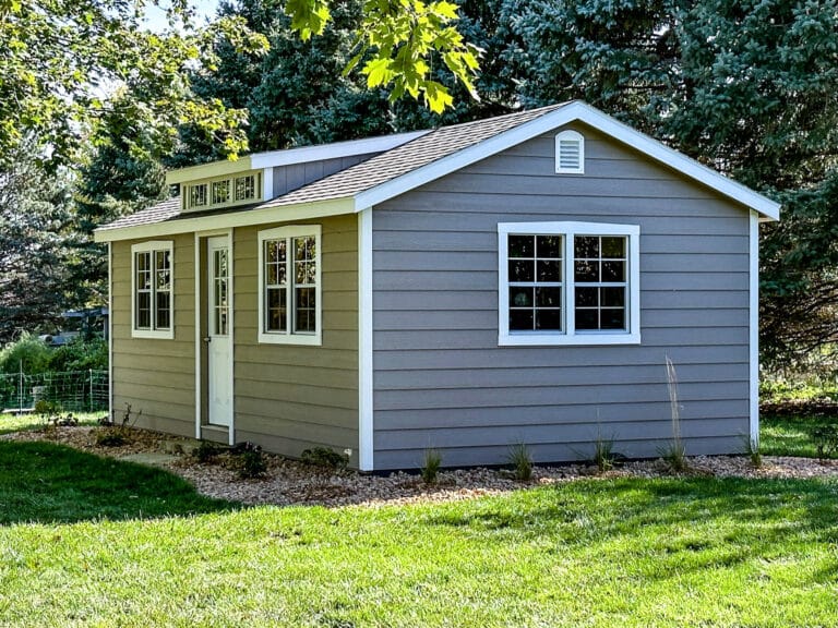 Light gray Southern Classic shed with white trim, multiple tall windows, a single entry door, and a transom dormer, set in a shaded grassy yard.