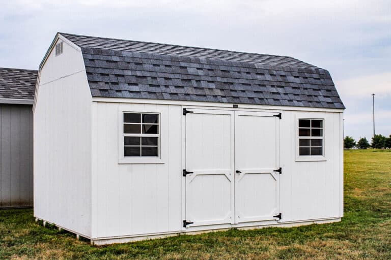 White gambrel-roof shed with double doors and two grid windows, situated on a grassy lot under a cloudy sky.