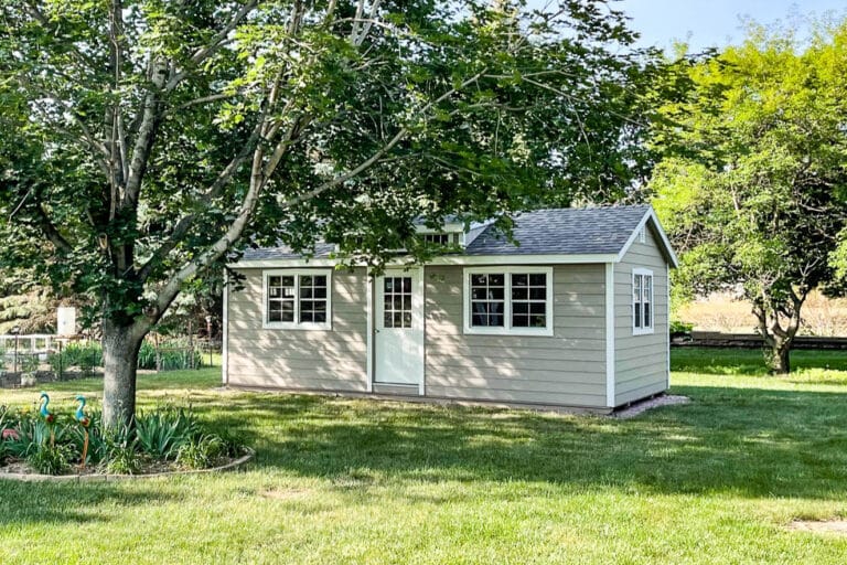 Light gray Southern Classic shed with white trim and multiple windows, shaded beneath large trees in a landscaped backyard.