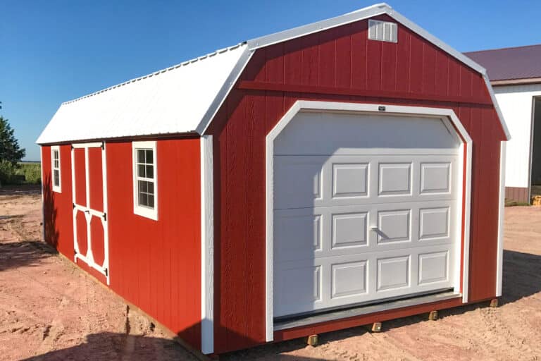 Another view of one of our red painted wood lofted 16x48 - Red barn-style storage shed with a white roof and garage door, placed on a sandy ground under a clear blue sky